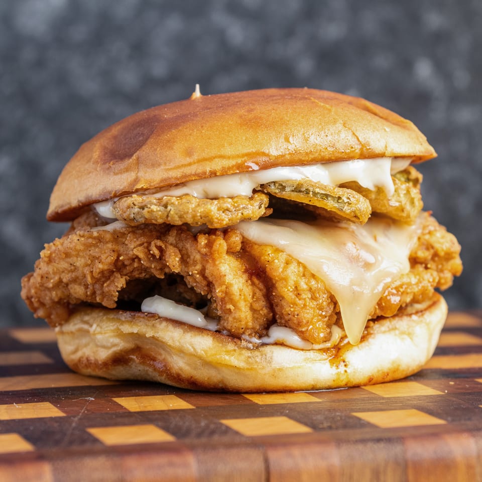 Close-up of a crispy fried chicken sandwich with melted cheese, fried jalapeños, and creamy sauce on a wooden board.