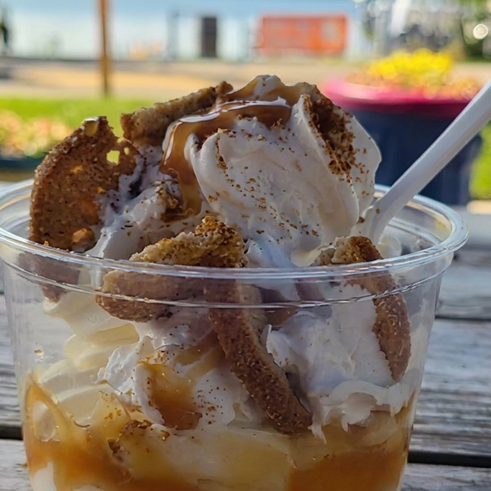 Vanilla soft-serve sundae topped with caramel, cookie pieces, and cinnamon, served in a clear cup with a spoon outdoors.
