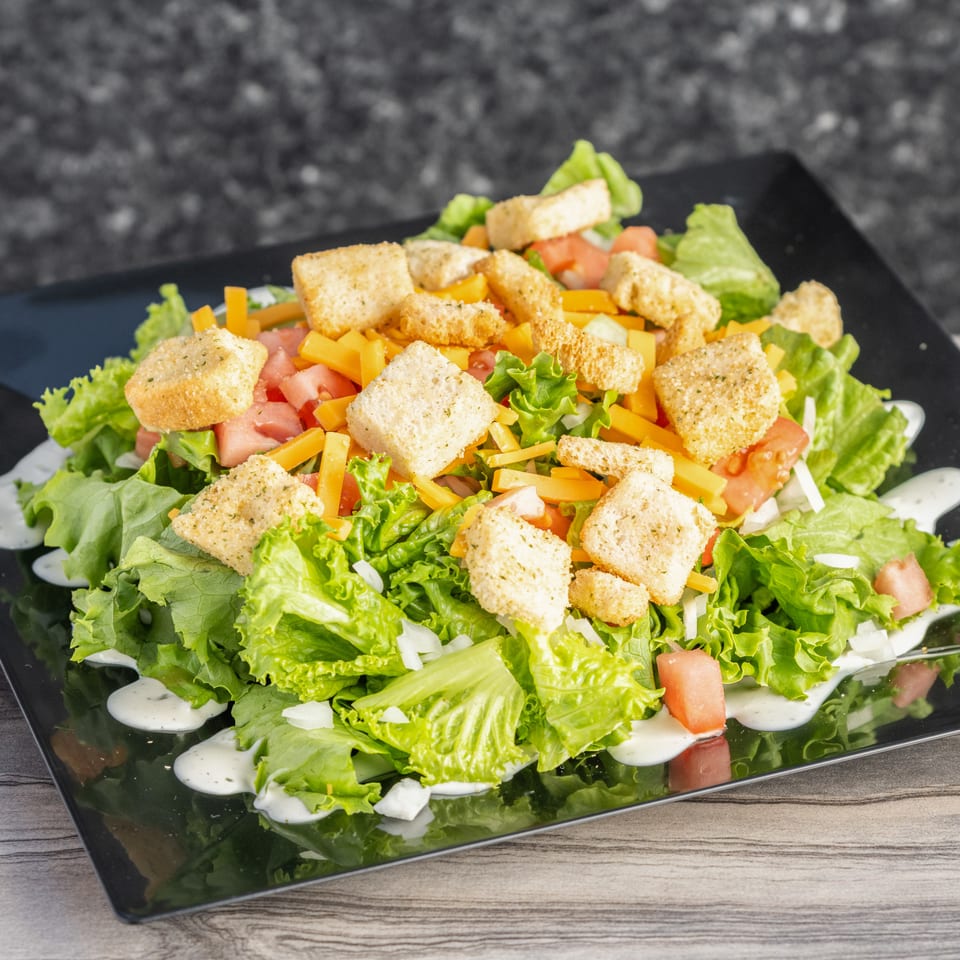 Fresh green salad with tomatoes, shredded cheese, croutons, and creamy dressing served on a black square plate.