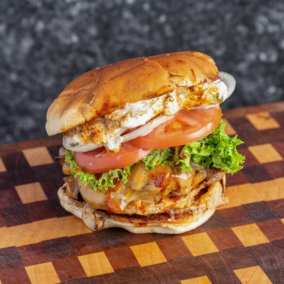 Stacked cheeseburger with grilled patties, creamy sauce, tomato, onion, and lettuce on a checkered wooden board.