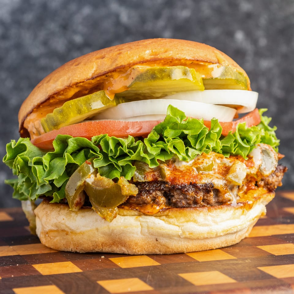 Close-up cheeseburger topped with lettuce, tomato, onion, pickles, peppers, and sauce on a checkered wooden board.