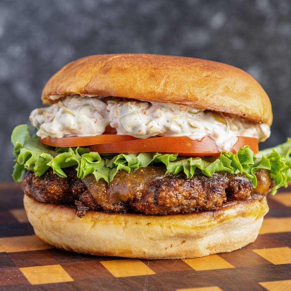Close-up of a loaded burger with a toasted bun, creamy coleslaw, tomato, lettuce, and a grilled beef patty on a checkered board.